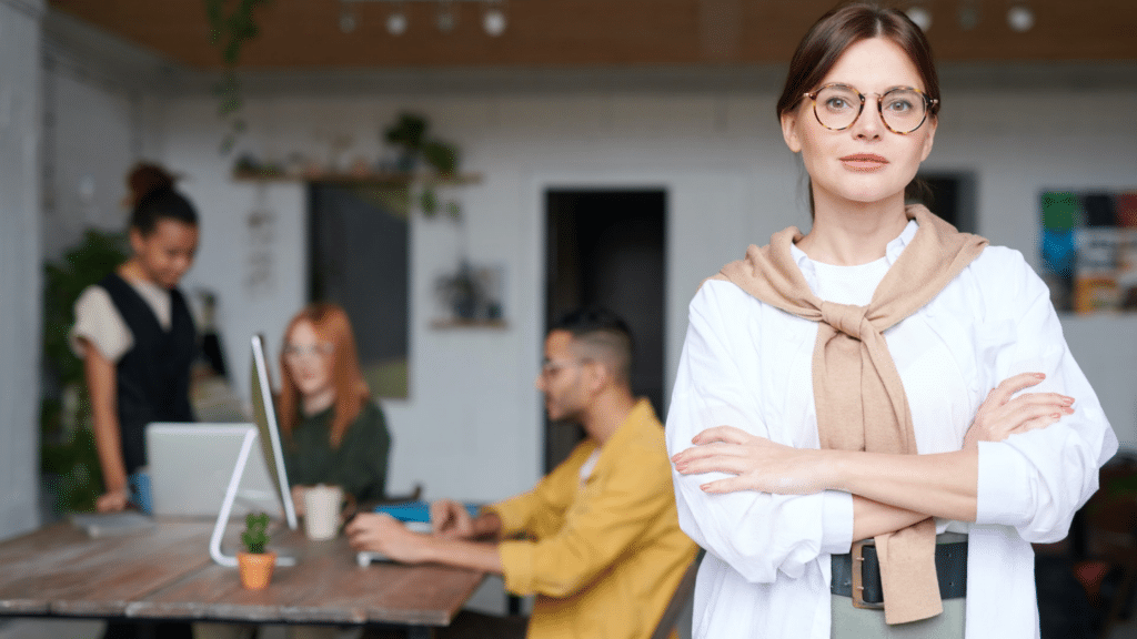 Une femme confiante posant les bras croisés au premier plan, avec une équipe collaborative travaillant en arrière-plan dans un bureau moderne